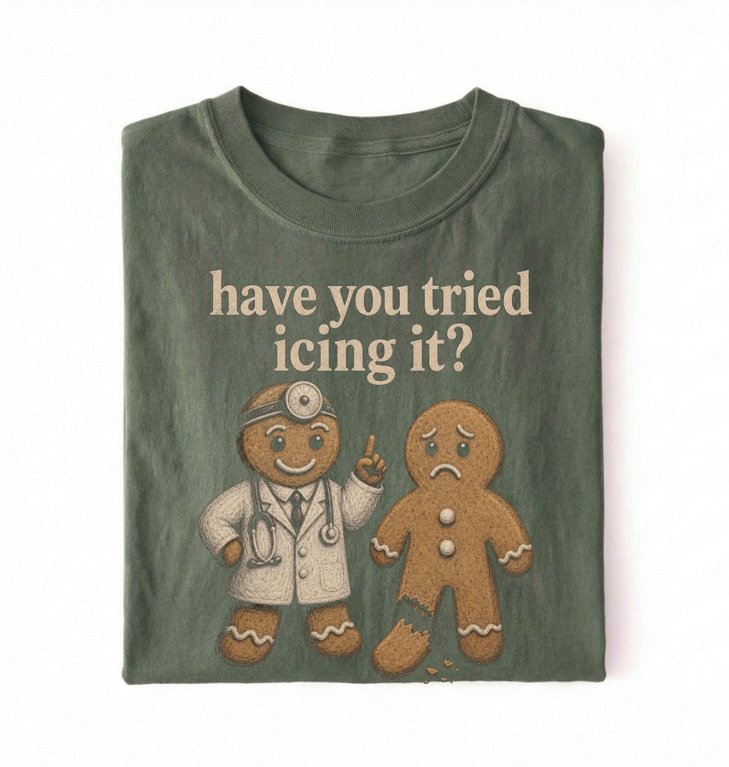 A folded dark olive green t-shirt on a white background, featuring a graphic of a gingerbread doctor and a sad gingerbread man with the text 'have you tried icing it?'.