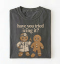 A folded dark gray t-shirt with a graphic print of two gingerbread men, one dressed as a doctor and the other sad, with the text 'have you tried icing it?' printed above them, on a white background.