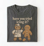 A folded dark gray t-shirt with a graphic print of two gingerbread men, one dressed as a doctor and the other sad, with the text 'have you tried icing it?' printed above them, on a white background.
