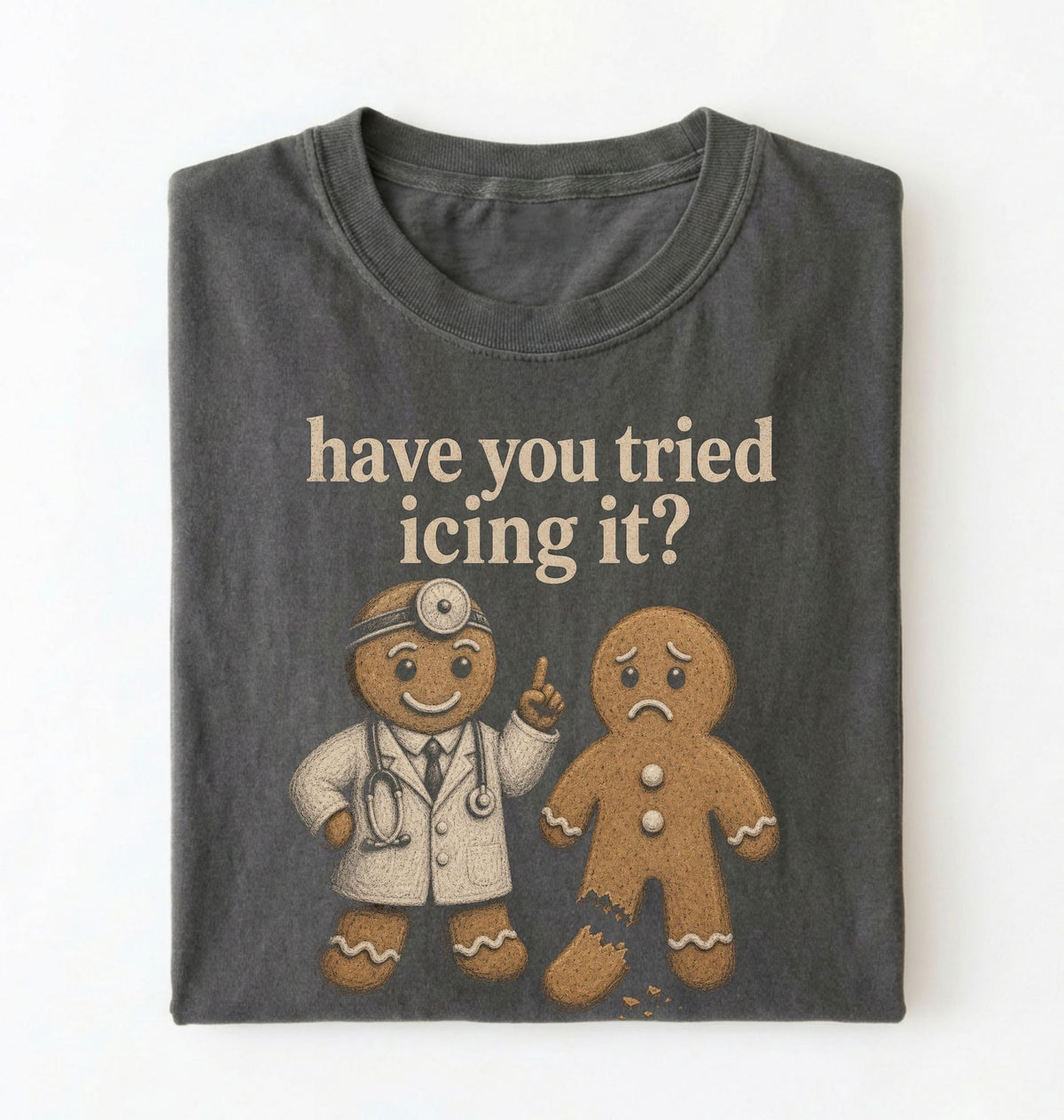 A folded dark gray t-shirt with a graphic print of two gingerbread men, one dressed as a doctor and the other sad, with the text 'have you tried icing it?' printed above them, on a white background.