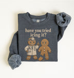 A folded dark heather gray sweatshirt with rolled sleeves, featuring a graphic of gingerbread characters and the text 'have you tried icing it?' on a plain off-white background.