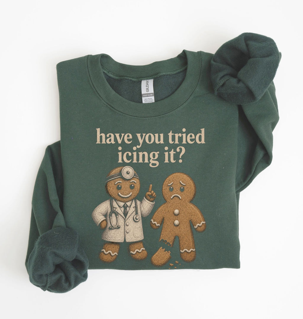 A dark green crewneck sweatshirt with a graphic print of a gingerbread doctor and the text 'have you tried icing it?' in beige. The sweatshirt is styled flat on a white background with rolled cuffs and a matching scrunchie.