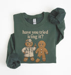 A dark green crewneck sweatshirt with a graphic print of a gingerbread doctor and the text 'have you tried icing it?' in beige. The sweatshirt is styled flat on a white background with rolled cuffs and a matching scrunchie.