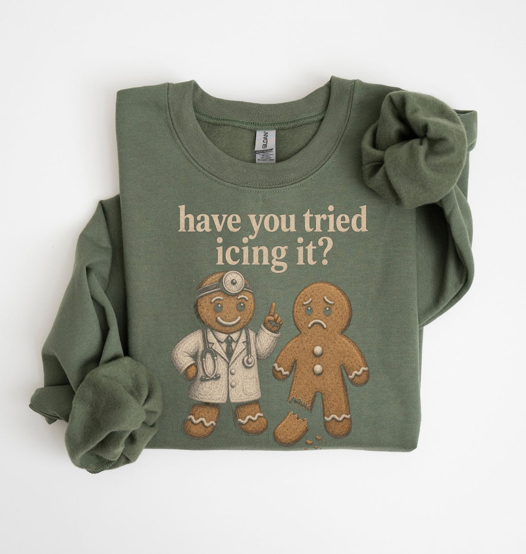 An olive green long-sleeve t-shirt with a graphic print of a gingerbread doctor and a sad gingerbread man, accompanied by the text 'have you tried icing it?'. The shirt is styled with a large scrunchie on one sleeve and is photographed flat against a white background.