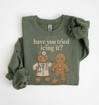 An olive green long-sleeve t-shirt with a graphic print of a gingerbread doctor and a sad gingerbread man, accompanied by the text 'have you tried icing it?'. The shirt is styled with a large scrunchie on one sleeve and is photographed flat against a white background.