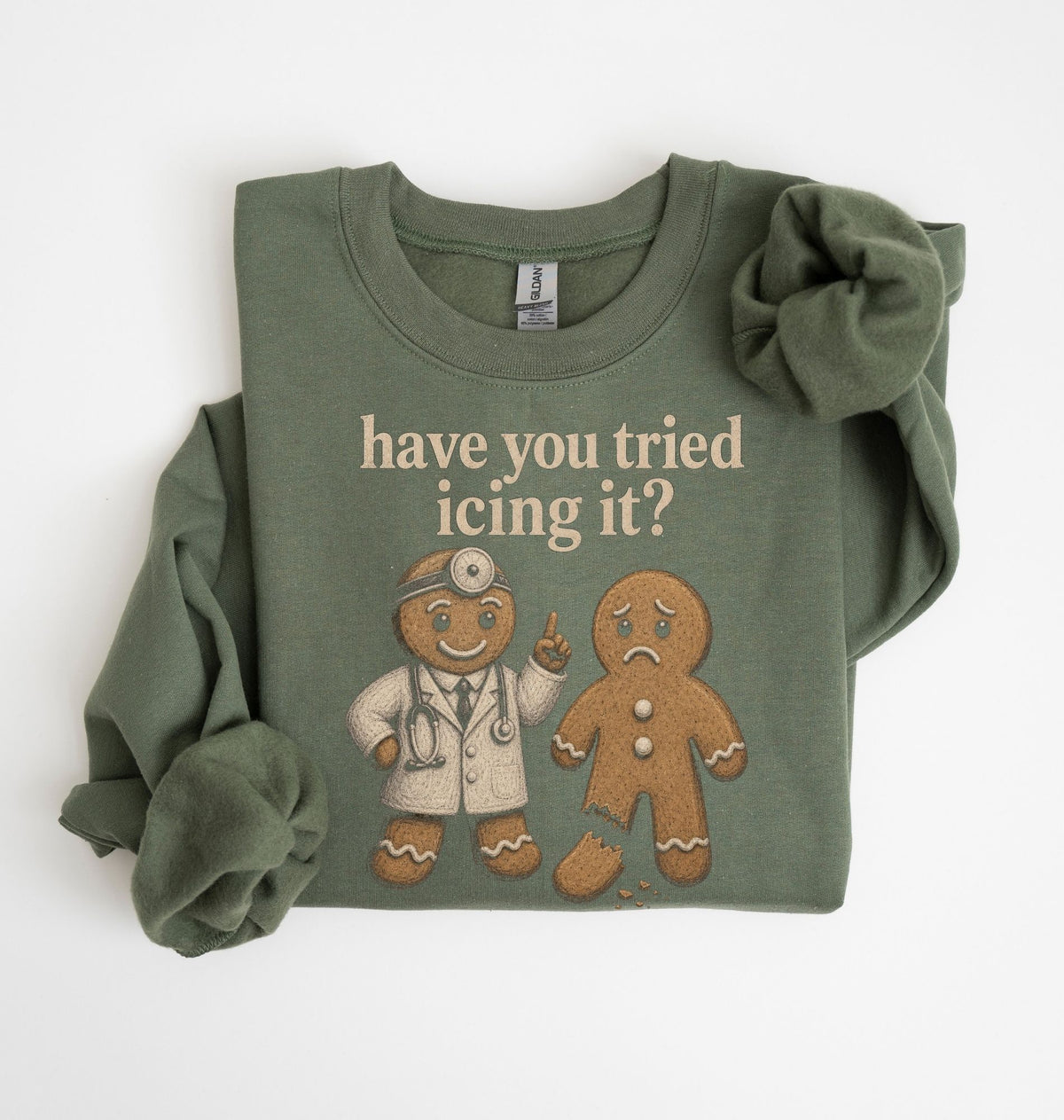 An olive green long-sleeve t-shirt with a graphic print of a gingerbread doctor and a sad gingerbread man, accompanied by the text 'have you tried icing it?'. The shirt is styled with a large scrunchie on one sleeve and is photographed flat against a white background.