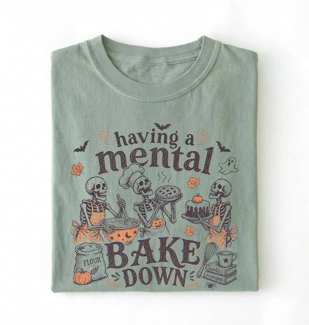 A folded sage green t-shirt with a Halloween-themed graphic print that reads 'having a mental BAKE DOWN' featuring cartoon skeletons baking, set against a plain white background.