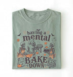 A folded sage green t-shirt with a Halloween-themed graphic print that reads 'having a mental BAKE DOWN' featuring cartoon skeletons baking, set against a plain white background.