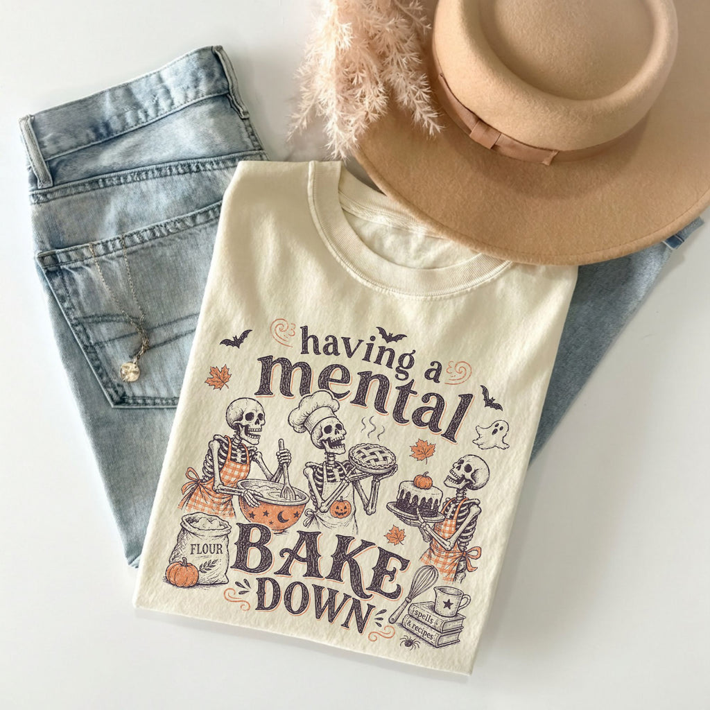 An off-white graphic t-shirt featuring a Halloween-themed baking skeleton design with the text 'having a mental BAKE DOWN', styled in a flat lay with light-wash jeans, a tan hat, and a fluffy beige feather on a white background.