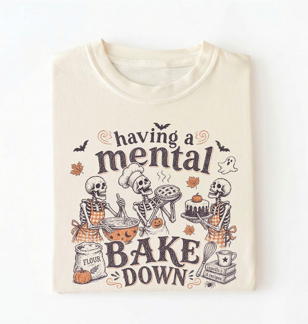 An off-white crewneck t-shirt featuring a Halloween-themed graphic of three skeletons baking, with the text 'having a mental BAKE DOWN' in dark brown lettering.
