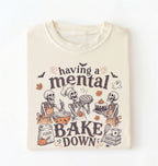 An off-white crewneck t-shirt featuring a Halloween-themed graphic of three skeletons baking, with the text 'having a mental BAKE DOWN' in dark brown lettering.