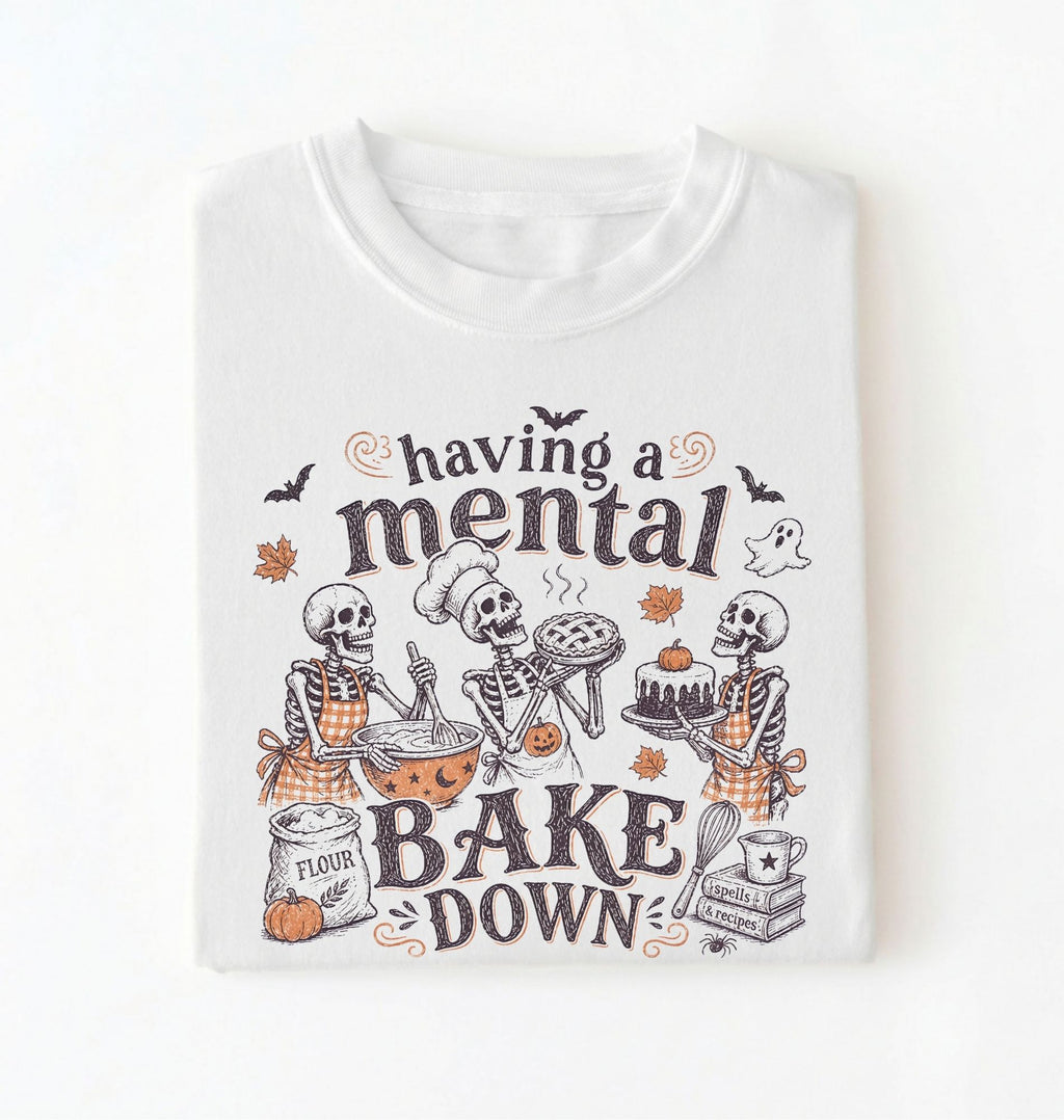 A white short-sleeve crewneck t-shirt featuring a Halloween-themed graphic print of three skeleton bakers with the text 'having a mental BAKE DOWN'. The graphic includes orange accents, bats, a ghost, and baking ingredients, shown on a clean white background.