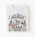 A white short-sleeve crewneck t-shirt featuring a Halloween-themed graphic print of three skeleton bakers with the text 'having a mental BAKE DOWN'. The graphic includes orange accents, bats, a ghost, and baking ingredients, shown on a clean white background.