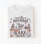 A white short-sleeve crewneck t-shirt featuring a Halloween-themed graphic print of three skeleton bakers with the text 'having a mental BAKE DOWN'. The graphic includes orange accents, bats, a ghost, and baking ingredients, shown on a clean white background.