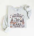A light heather gray long-sleeve sweatshirt with a Halloween-themed graphic print featuring cartoon skeletons baking and the text 'having a mental BAKE DOWN'. The sweatshirt is styled flat on a warm off-white background with one sleeve rolled up.