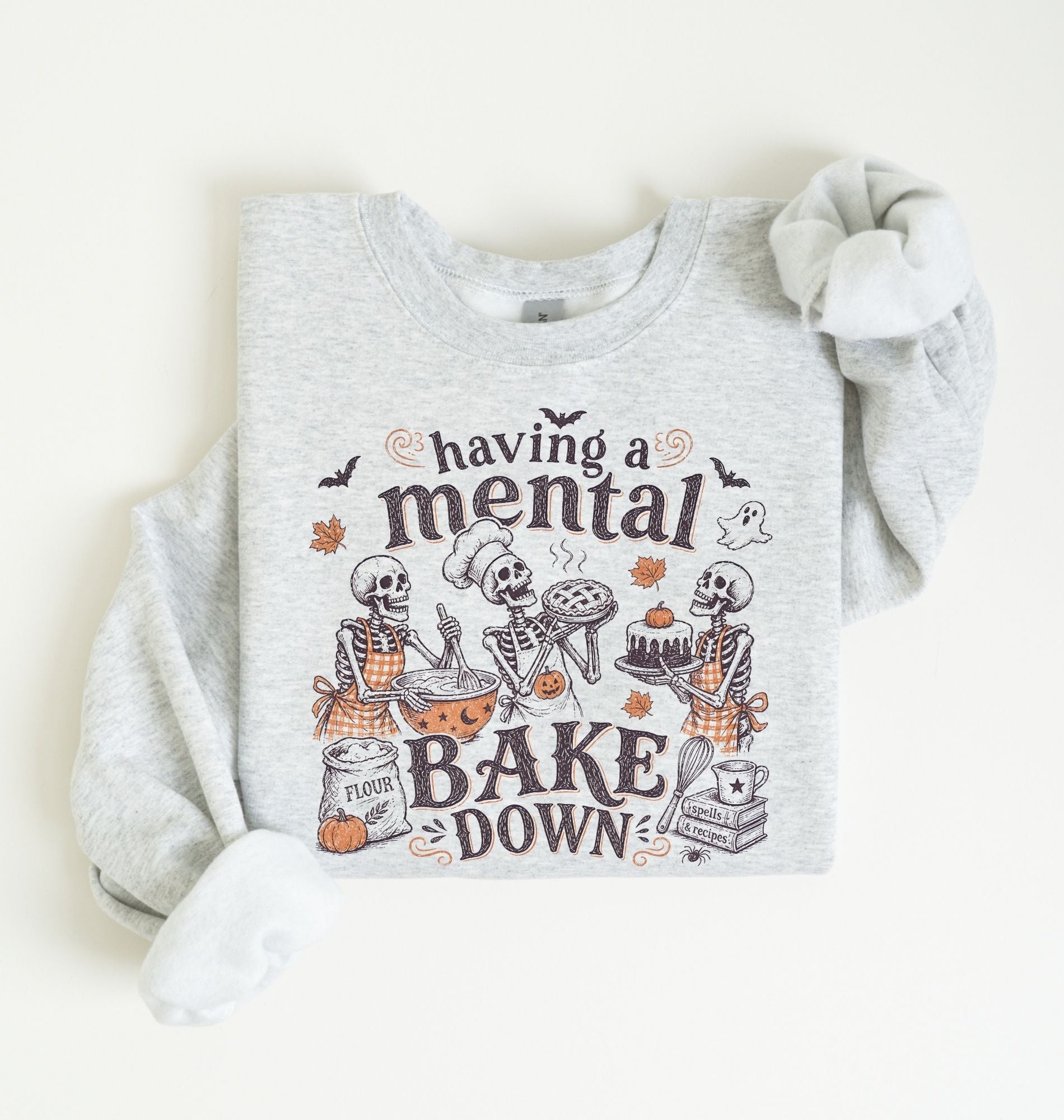 A light heather gray long-sleeve sweatshirt with a Halloween-themed graphic print featuring cartoon skeletons baking and the text 'having a mental BAKE DOWN'. The sweatshirt is styled flat on a warm off-white background with one sleeve rolled up.