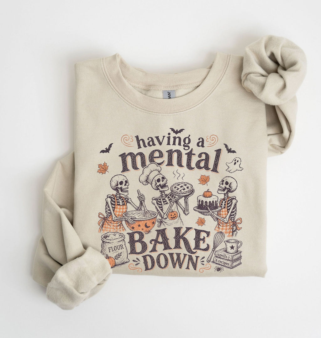 A beige crewneck sweatshirt with a Halloween-themed graphic print that reads 'having a mental BAKE DOWN' and features cartoon skeletons baking. The sweatshirt is styled flat on a white background with one sleeve rolled up and the other cuff resting beside it.