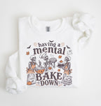 A white long-sleeve crewneck sweatshirt with a graphic print of skeleton bakers baking treats, accompanied by the text 'having a mental BAKE DOWN'.