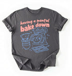 Flat lay of a Having a Mental Bake Down baking shirt with mixer graphic on brown t-shirt on a ivory background