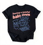 Flat lay of a Having a Mental Bake Down baking shirt with mixer graphic on black t-shirt on a ivory background