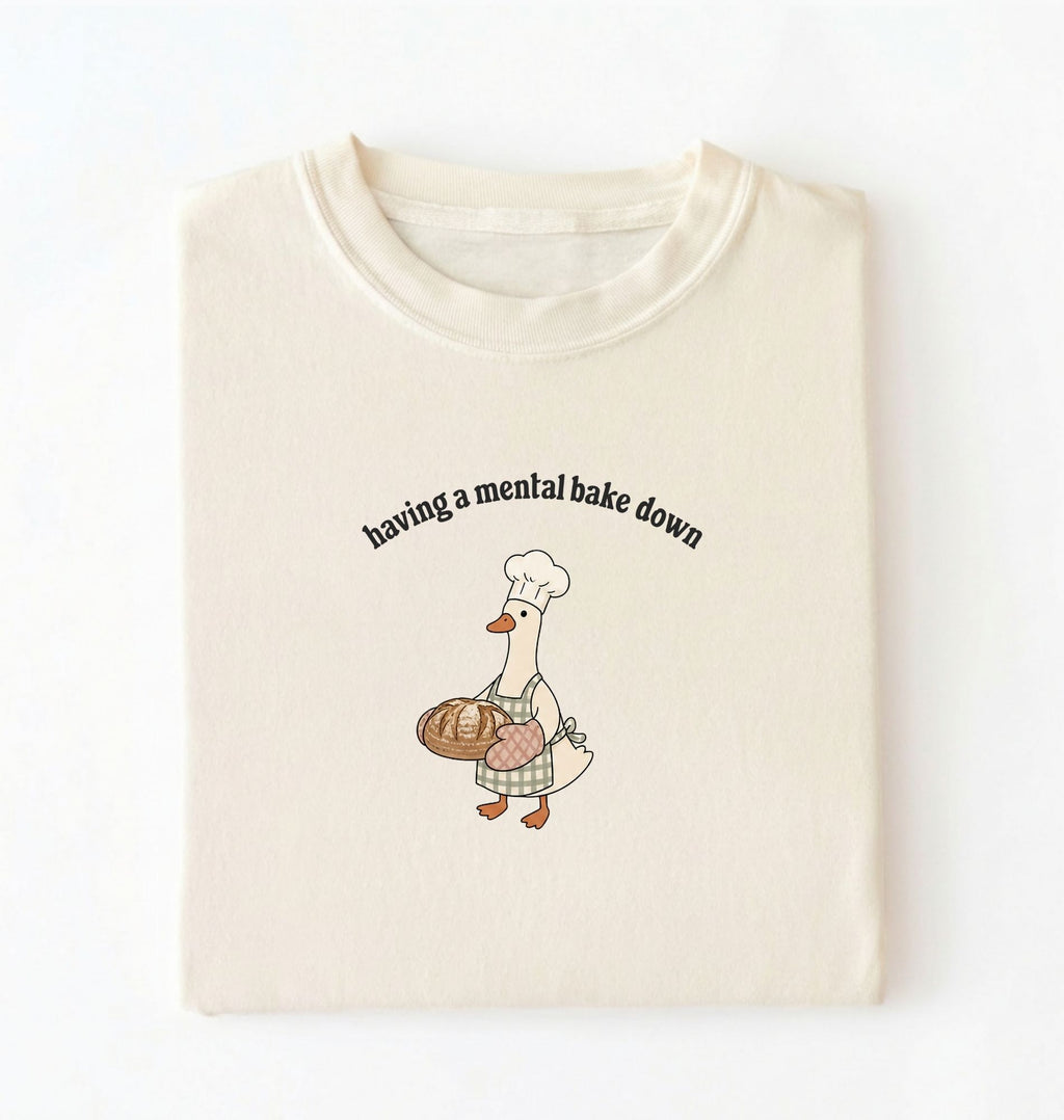 Flat lay of a cream Funny Having a Mental Bake Down shirt with baker goose holding artisan bread graphic on a white background 