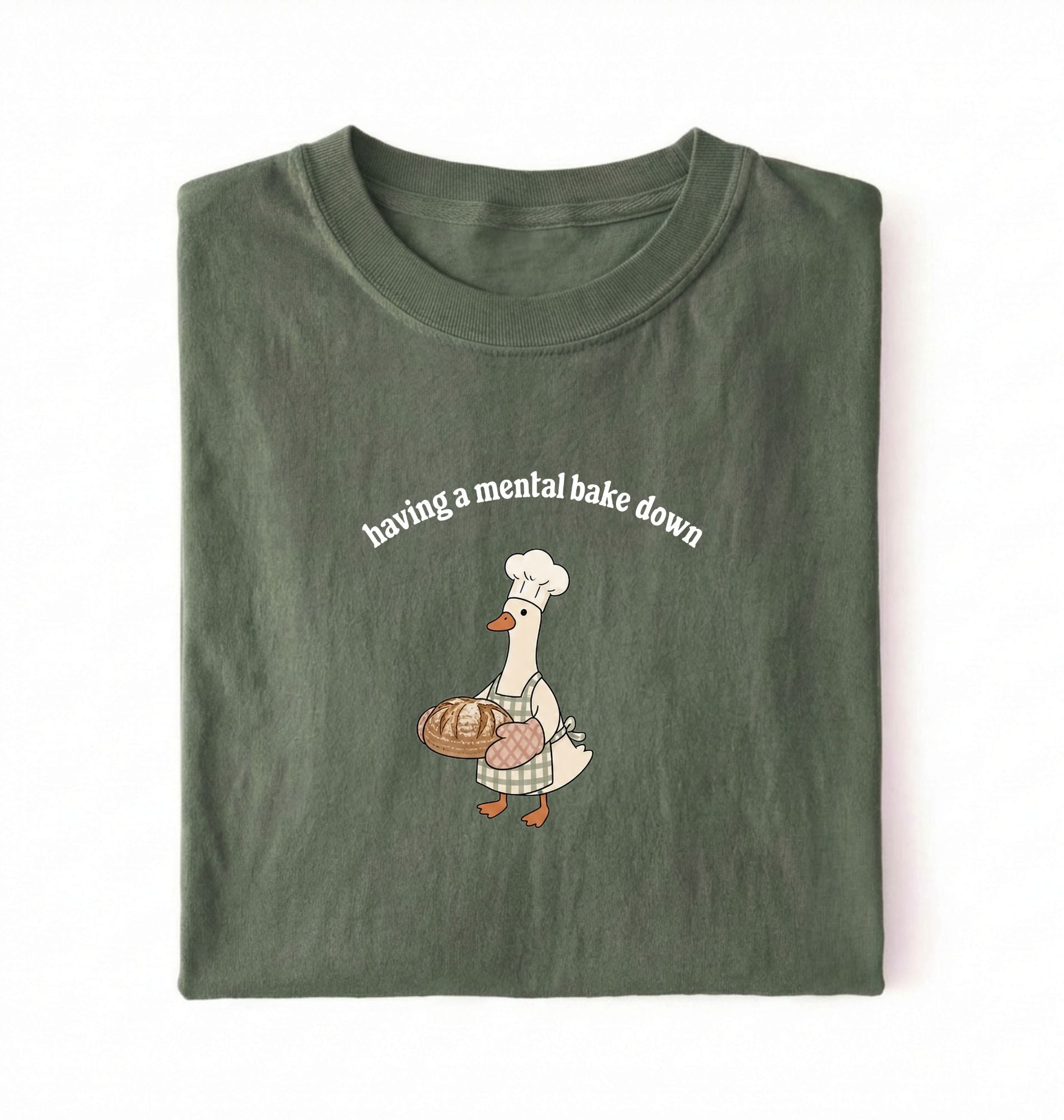 Flat lay of a green Funny Having a Mental Bake Down shirt with baker goose holding artisan bread graphic on a white background 