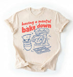 Flat lay of a Having a Mental Bake Down baking shirt with mixer graphic on brown t-shirt on a ivory background