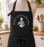 A black kitchen apron displayed on a beige mannequin torso in a bright kitchen setting. The apron has a white printed graphic of a skeleton holding a bowl, with the text 'HAVING A MENTAL BAKE DOWN' arched above it.