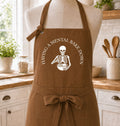 A brown apron displayed on a mannequin torso in a bright kitchen setting. The apron has a white printed graphic of a skeleton holding a mixing bowl, with the text 'HAVING A MENTAL BAKE DOWN' curved above it.