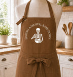 A brown apron displayed on a mannequin torso in a bright kitchen setting. The apron has a white printed graphic of a skeleton holding a mixing bowl, with the text 'HAVING A MENTAL BAKE DOWN' curved above it.