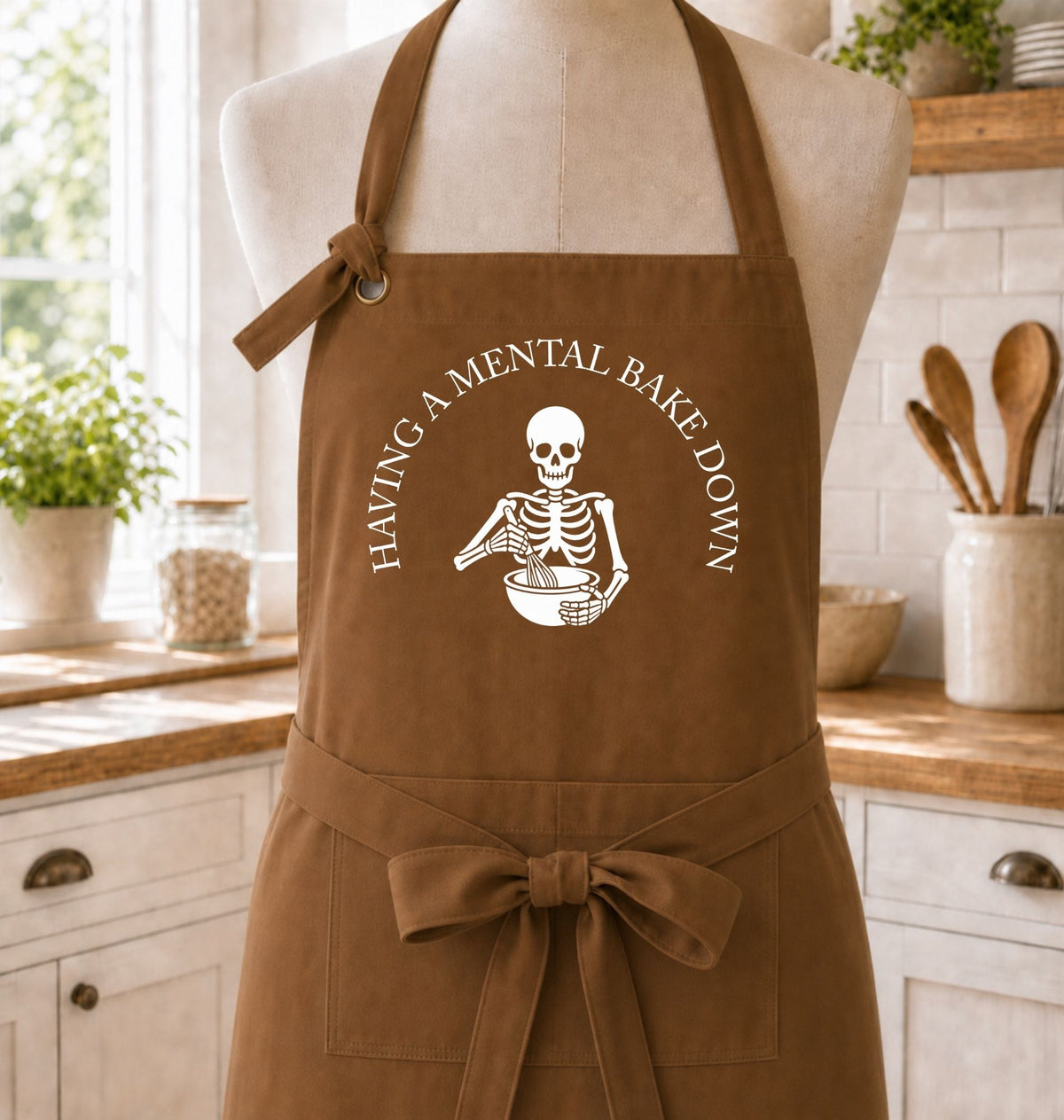 A brown apron displayed on a mannequin torso in a bright kitchen setting. The apron has a white printed graphic of a skeleton holding a mixing bowl, with the text 'HAVING A MENTAL BAKE DOWN' curved above it.