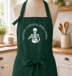 A dark green kitchen apron displayed on a dress form mannequin in a bright kitchen setting. The apron features a white graphic of a skeleton holding a bowl with the text 'HAVING A MENTAL BAKE DOWN' arched above it.