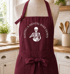 A burgundy kitchen apron with a white skeleton graphic and the text 'HAVING A MENTAL BAKE DOWN' displayed on a mannequin torso in a bright kitchen setting.