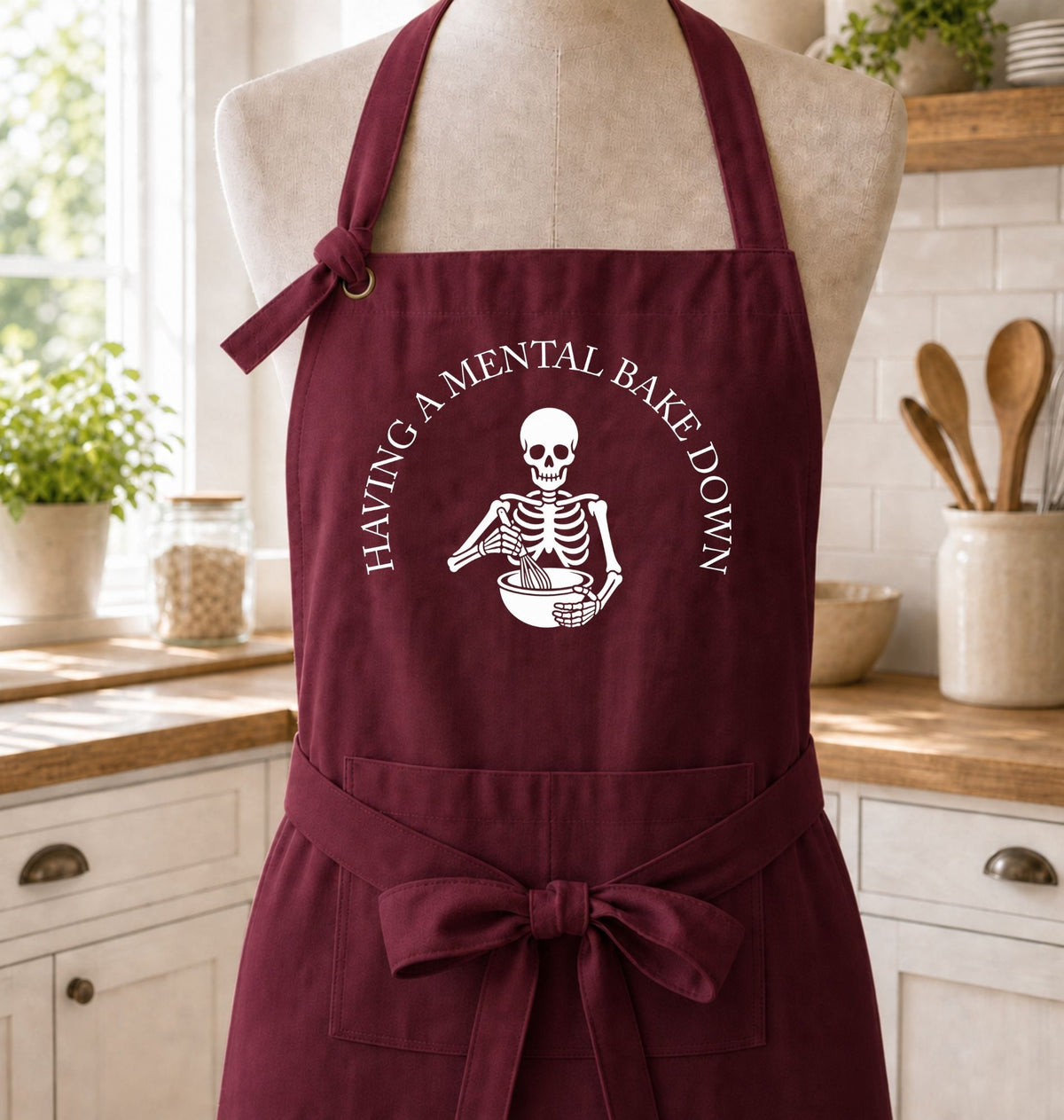 A burgundy kitchen apron with a white skeleton graphic and the text 'HAVING A MENTAL BAKE DOWN' displayed on a mannequin torso in a bright kitchen setting.