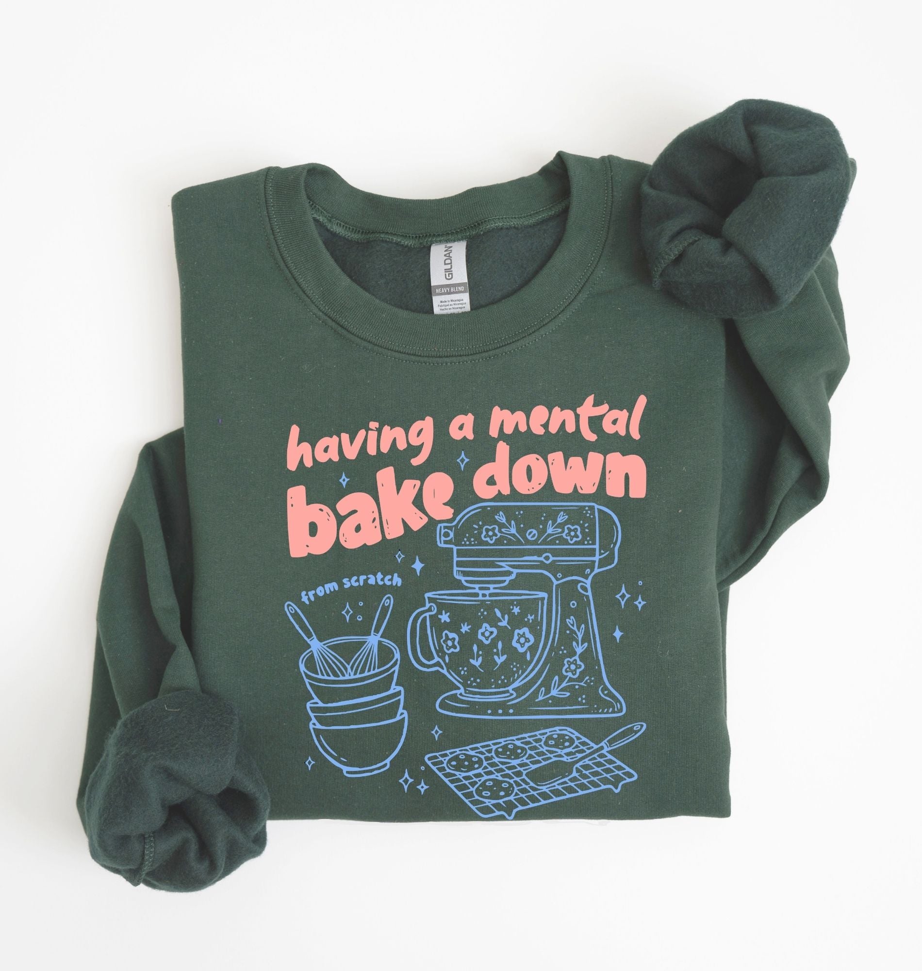 Flat lay of a forest green Funny Having a Mental Bake Down baking sweatshirt with mixer and baking tools graphic on a white background 