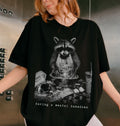 A woman wearing a black oversized graphic t-shirt with a black and white print of a raccoon eating pizza and the text 'Having a mental bakedown'.