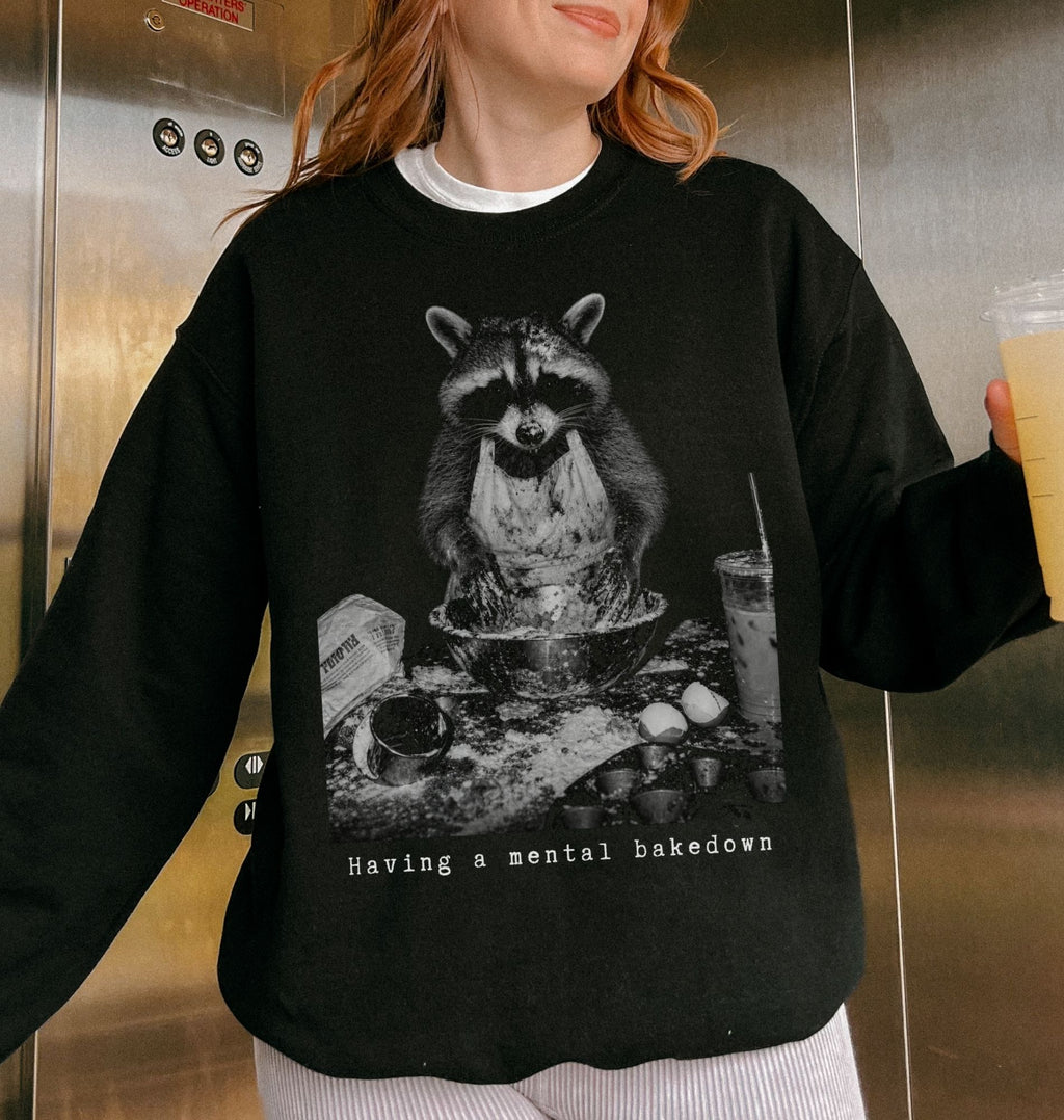 A person wearing a black crewneck sweatshirt with a large black-and-white graphic of a raccoon sitting at a table with the text 'Having a mental bakedown' below it. The person is standing in front of a metallic elevator wall and holding a drink.
