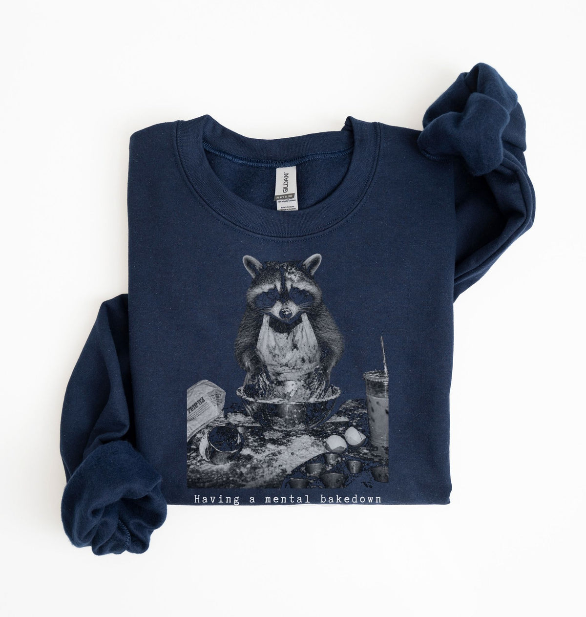 A navy blue sweatshirt with a graphic of a raccoon sitting in a messy bowl of food, with the text 'Having a mental breakdown' printed below it. The sweatshirt is laid flat on a white background with one sleeve folded over.