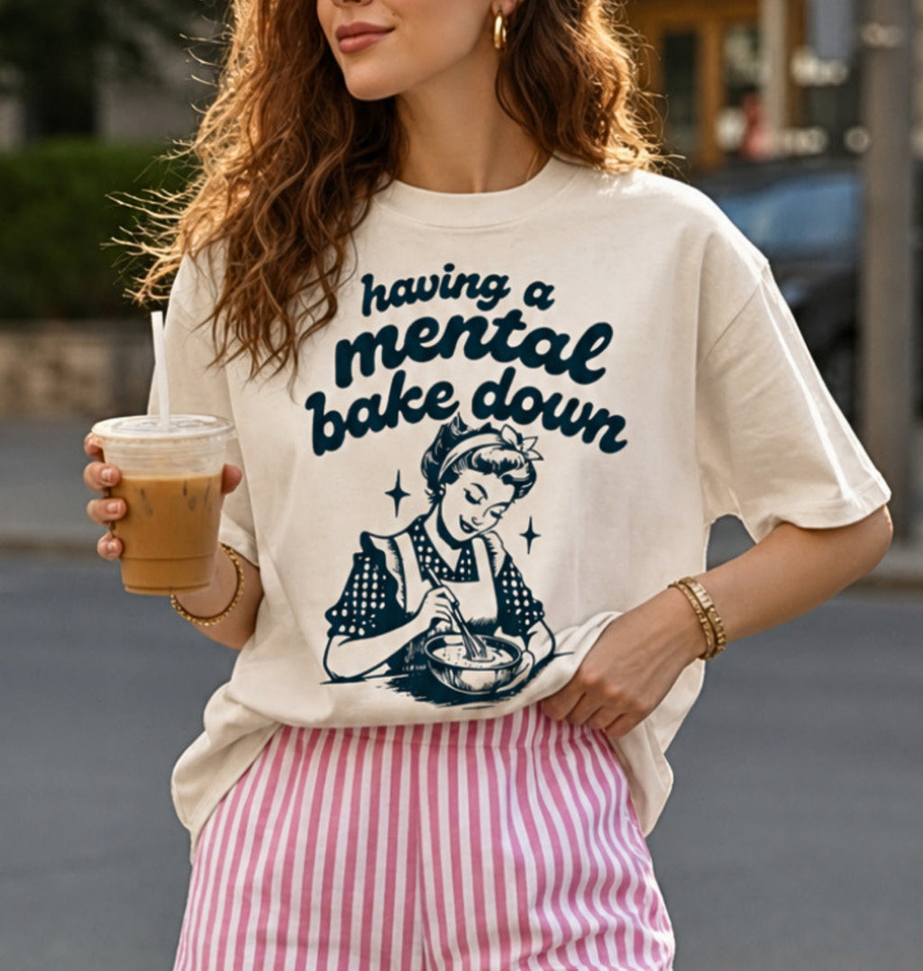 A woman wearing an oversized cream graphic t-shirt with the text 'having a mental bake down' and a retro illustration of a woman baking, paired with pink and white striped pants, holding an iced coffee in an outdoor setting.