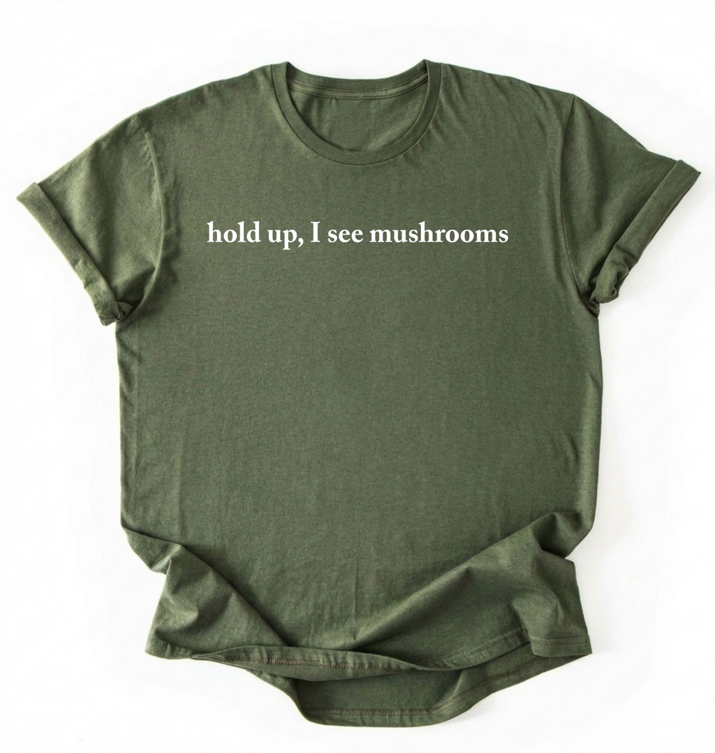 Flat lay of a moss green colored t-shirt on a white background featuring brown text that reads “hold up, I see mushrooms. 