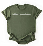 Flat lay of a moss green colored t-shirt on a white background featuring brown text that reads “hold up, I see mushrooms. 