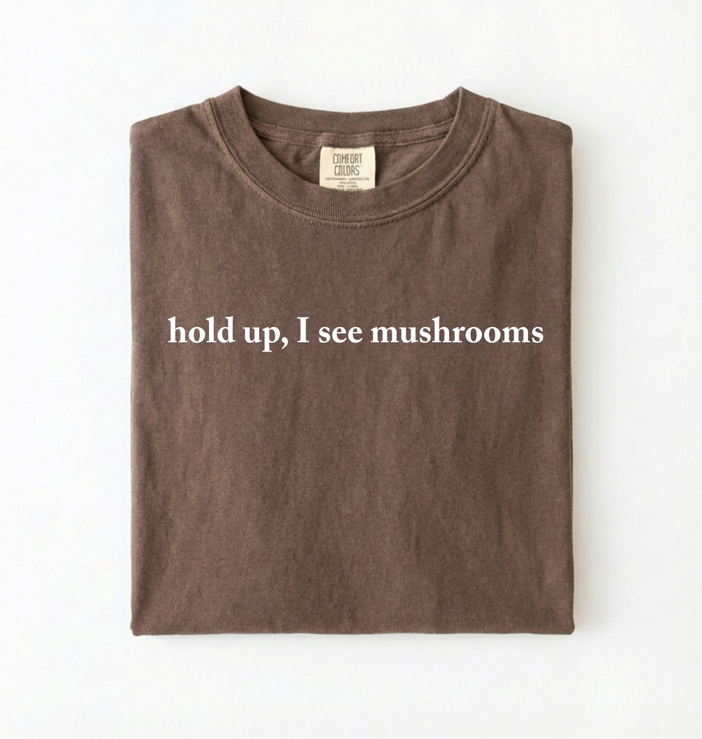 Flat lay of a espresso brown colored t-shirt on a white background featuring brown text that reads “hold up, I see mushrooms. 