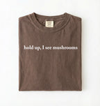 Flat lay of a espresso brown colored t-shirt on a white background featuring brown text that reads “hold up, I see mushrooms. 