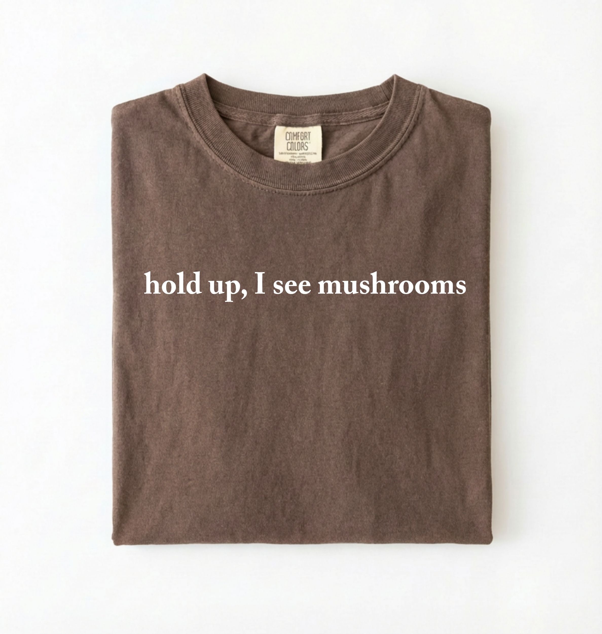 Flat lay of a espresso brown colored t-shirt on a white background featuring brown text that reads “hold up, I see mushrooms. 