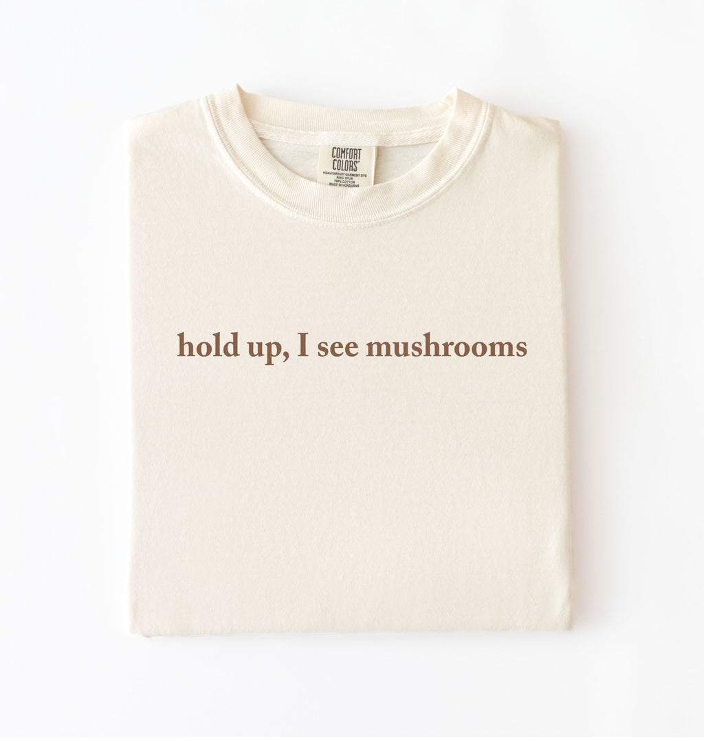 Flat lay of an ivory colored t-shirt on a white background featuring brown text that reads “hold up, I see mushrooms. 