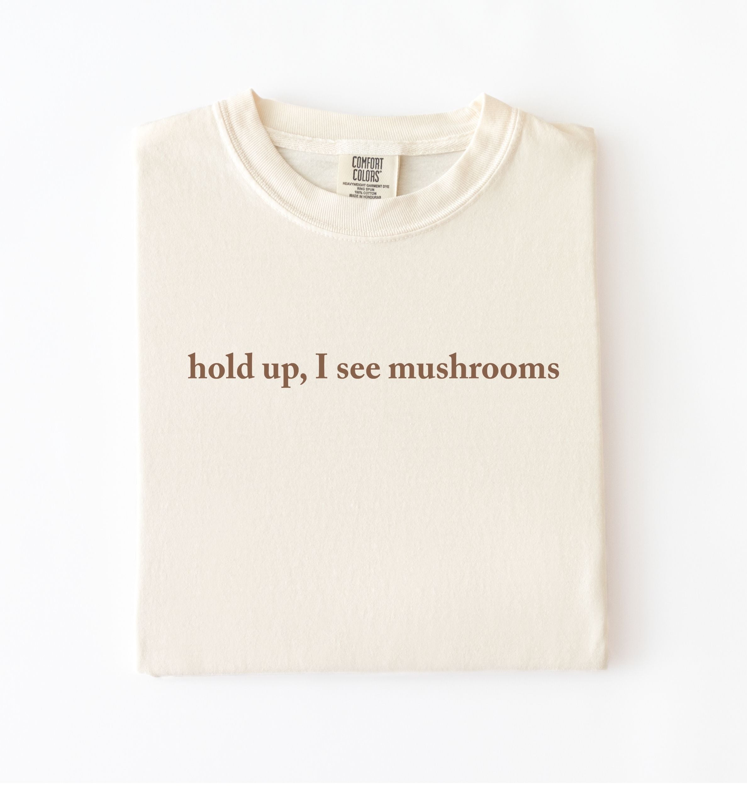 Flat lay of an ivory colored t-shirt on a white background featuring brown text that reads “hold up, I see mushrooms. 
