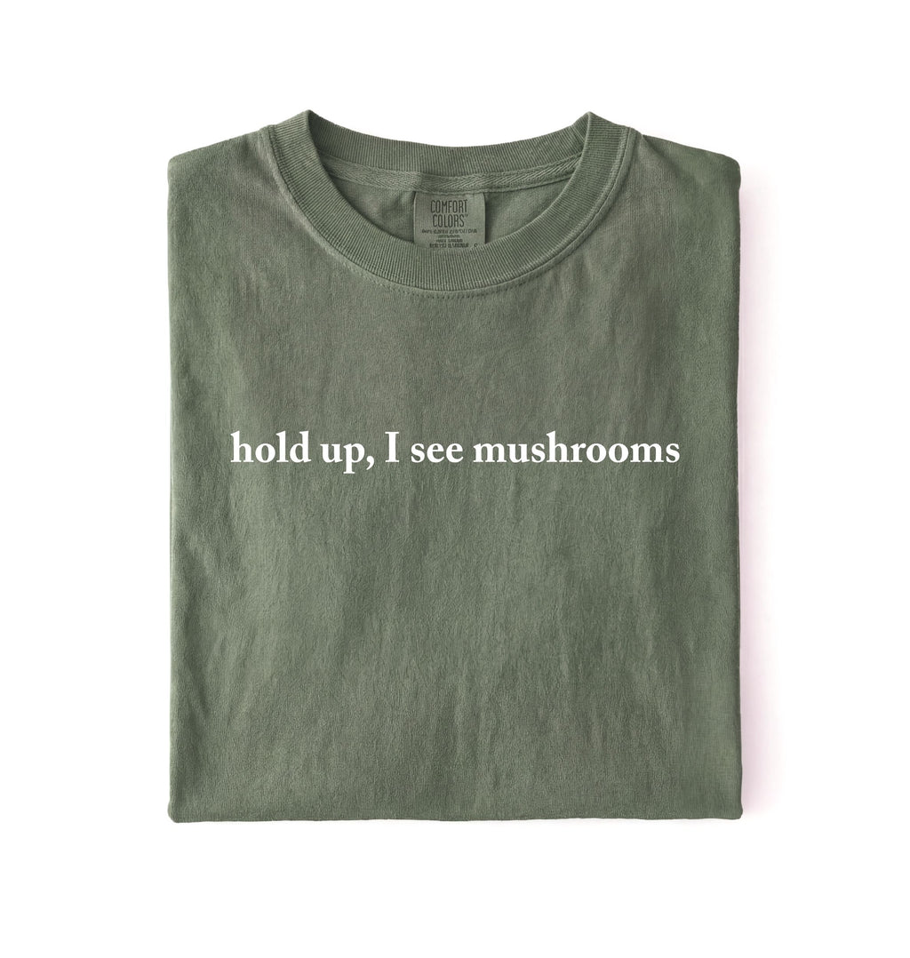 Flat lay of a moss green colored t-shirt on a white background featuring brown text that reads “hold up, I see mushrooms. 