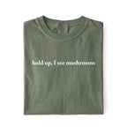Flat lay of a moss green colored t-shirt on a white background featuring brown text that reads “hold up, I see mushrooms. 