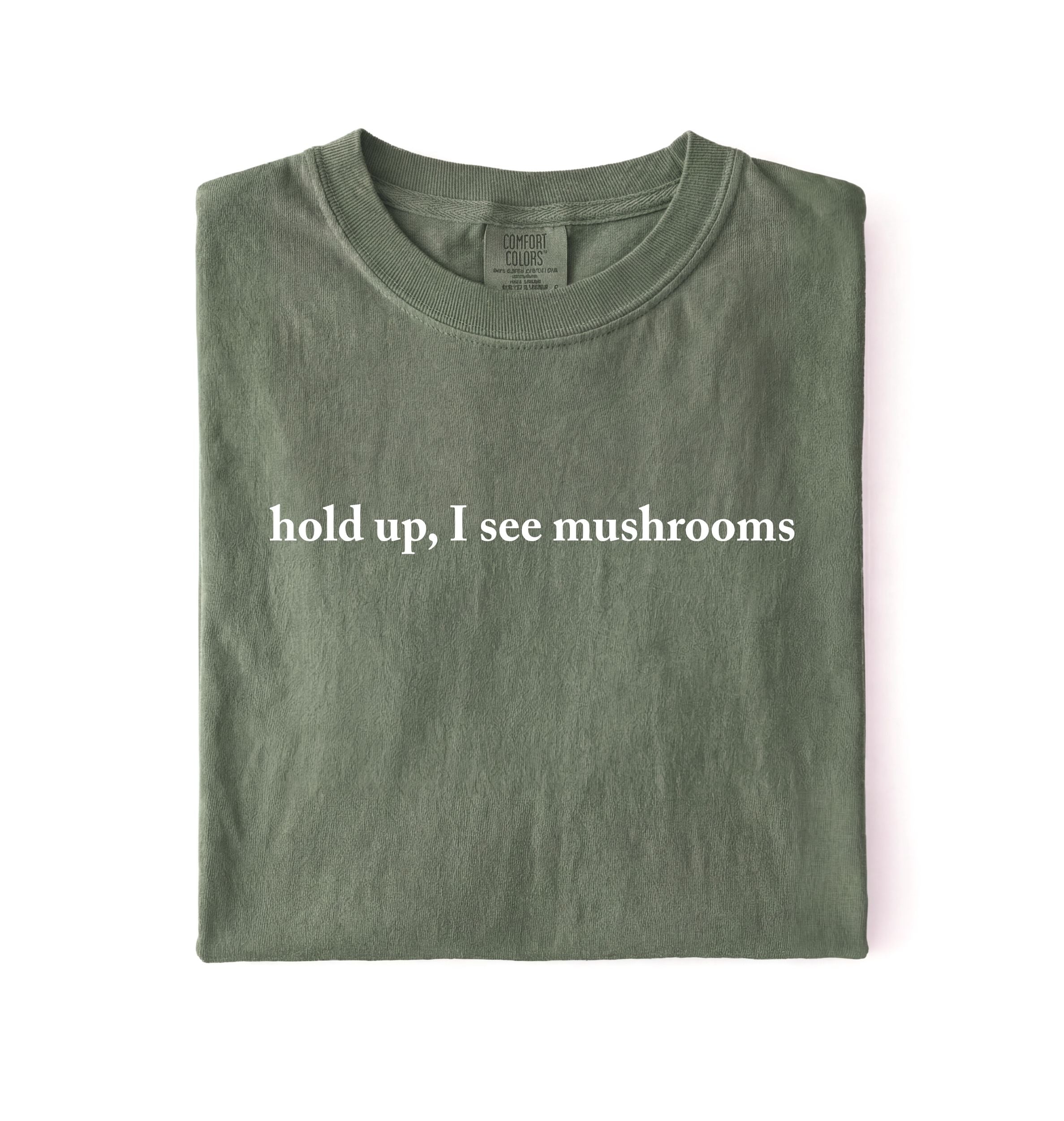 Flat lay of a moss green colored t-shirt on a white background featuring brown text that reads “hold up, I see mushrooms. 