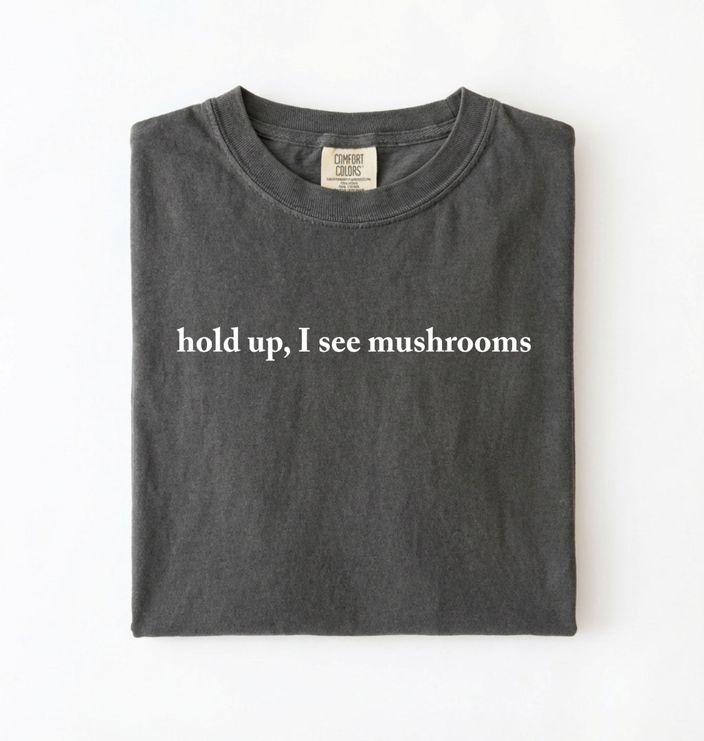 Flat lay of a pepper-colored t-shirt on a white background featuring brown text that reads “hold up, I see mushrooms. 